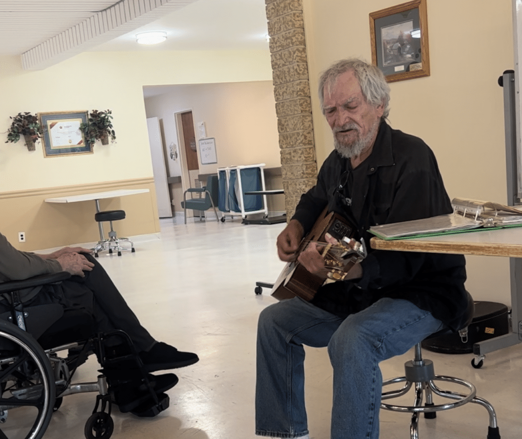 Older man plays guitar to someone in wheelchair.