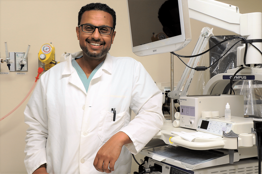 Doctor in lab coat smiles at camera and leans arm on piece of medical equipment.