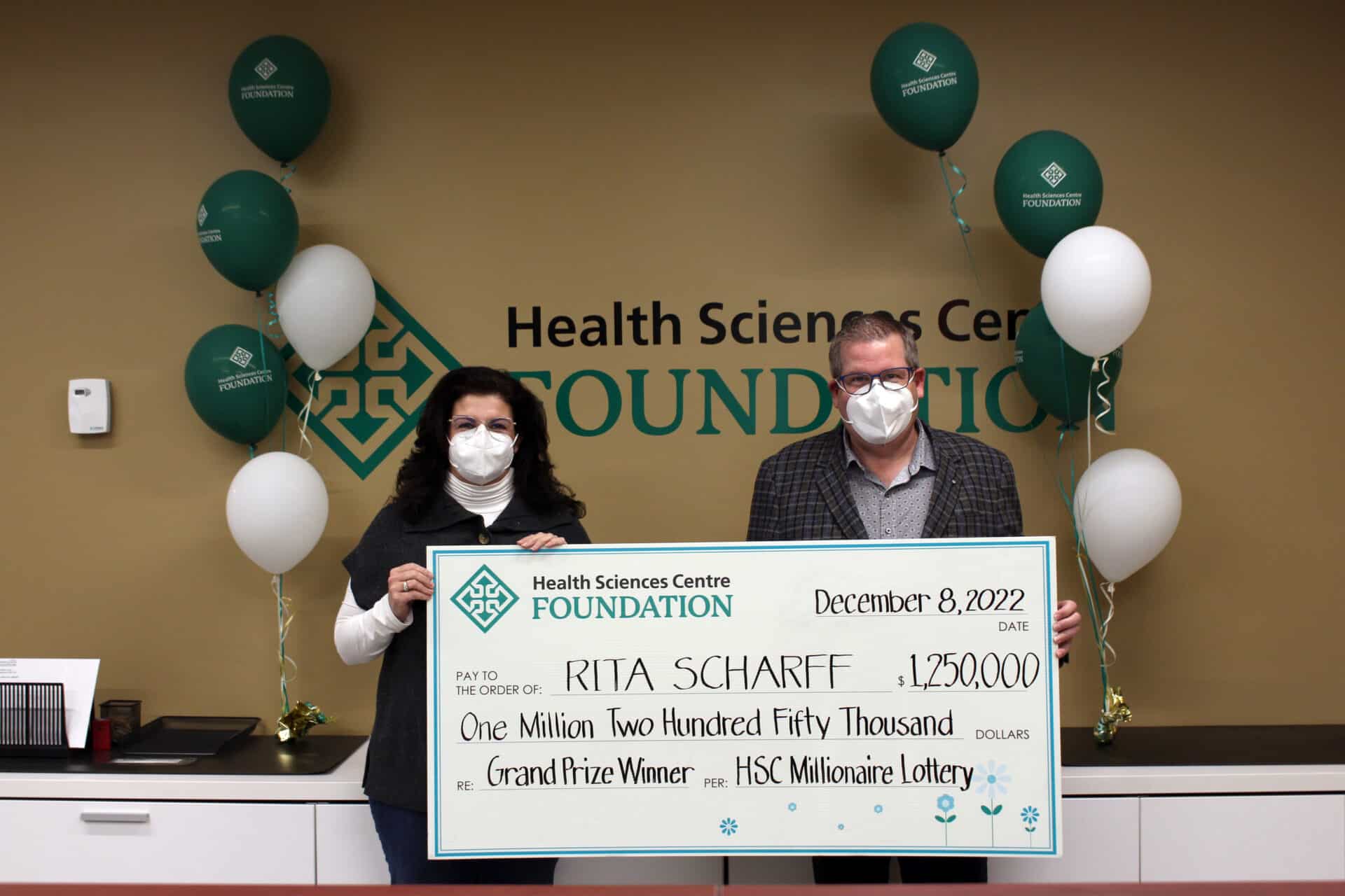 Woman with black curly hair, wearing a black shirt overtop a white turtleneck with glasses and a mask, holds up an oversized cheque with a man wearing a suit jacket, grey shirt, glasses, and mask.