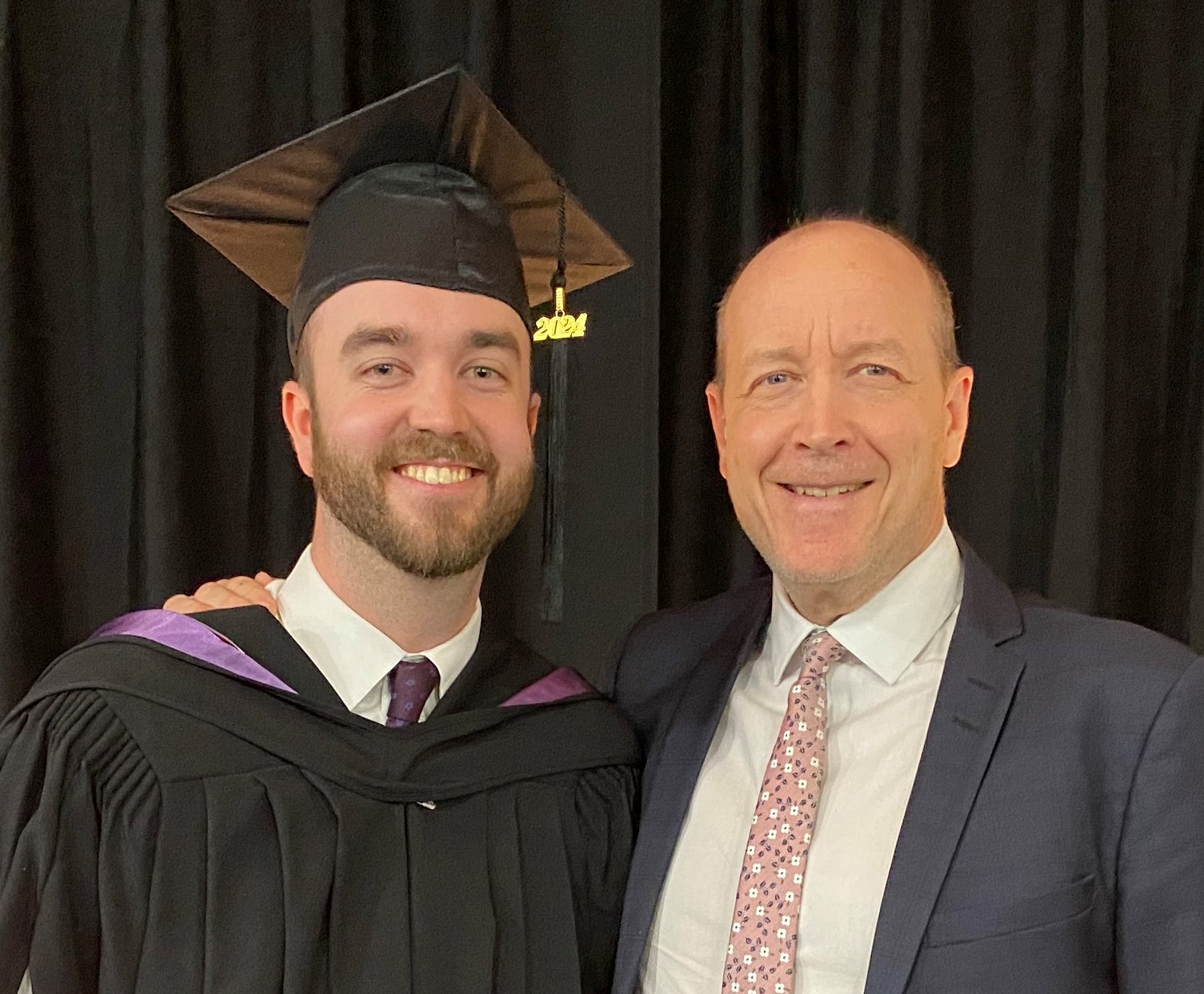 A younger man in a black graduation cap and gown smiles at the camera. A smiling older man in a blue suit and pink tie has his arm around the younger man's shoulders.