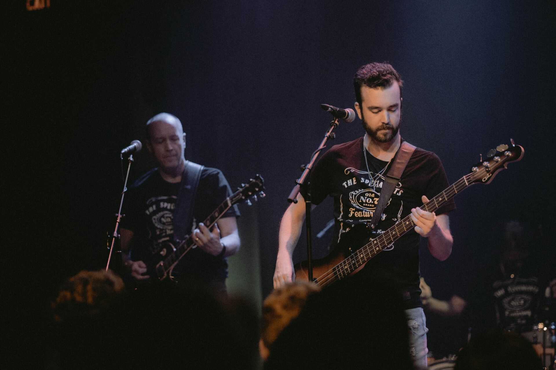 Two men standing on stage performing as part of a band. Both men are holding guitars and standing in front of microphones.