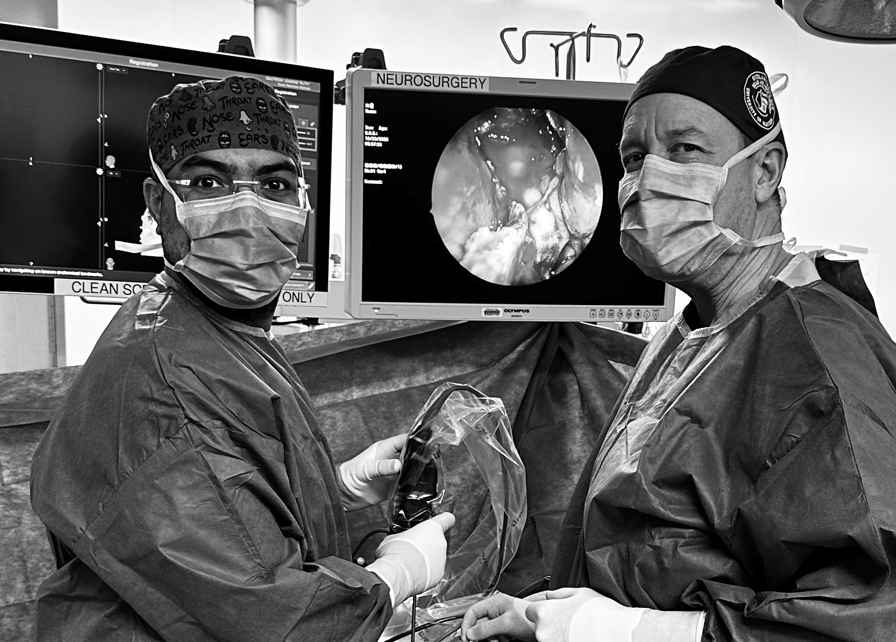 A black and white photo of two doctors performing surgery. The doctors are standing in front of two computer monitors labelled 'Neurosurgery'; the image on the computer screen shows a photo of the inside of a patient's skull.