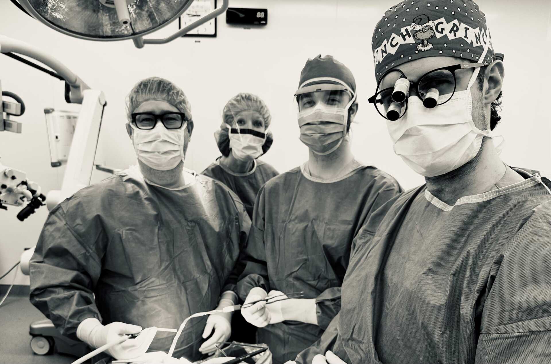 A sepia toned photo shows four doctors in surgical wear, including gowns, caps, and face masks, looking at the camera.
